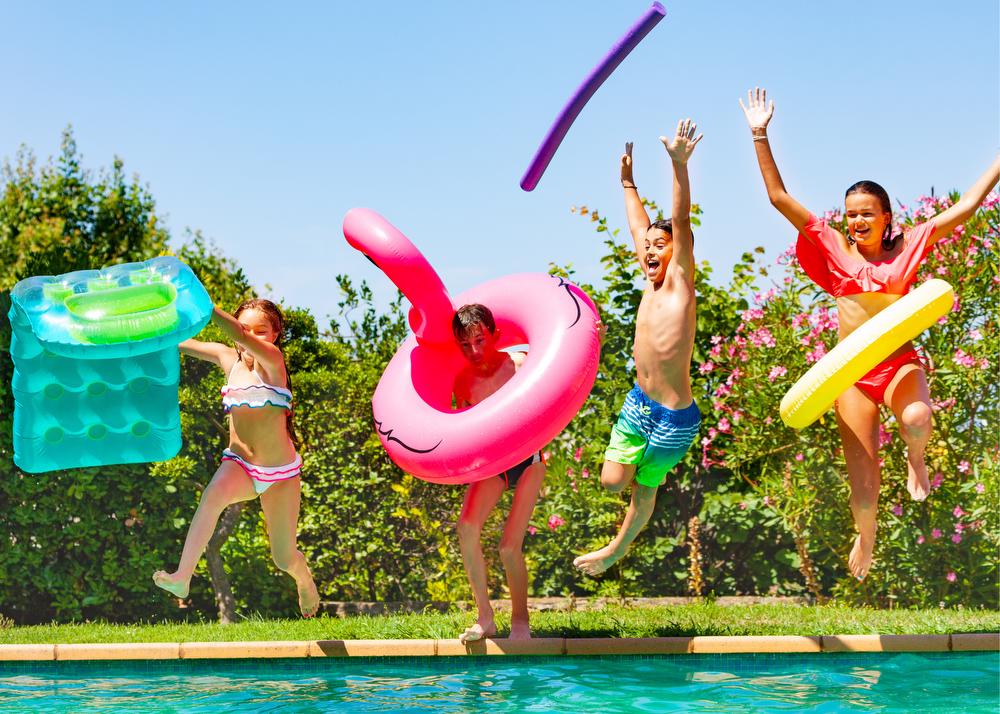 Four kids in bathing suits jumping off the side of a pool with inflatables