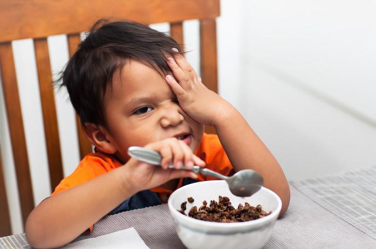 Young child eating cereal with one hand on his face and scowling