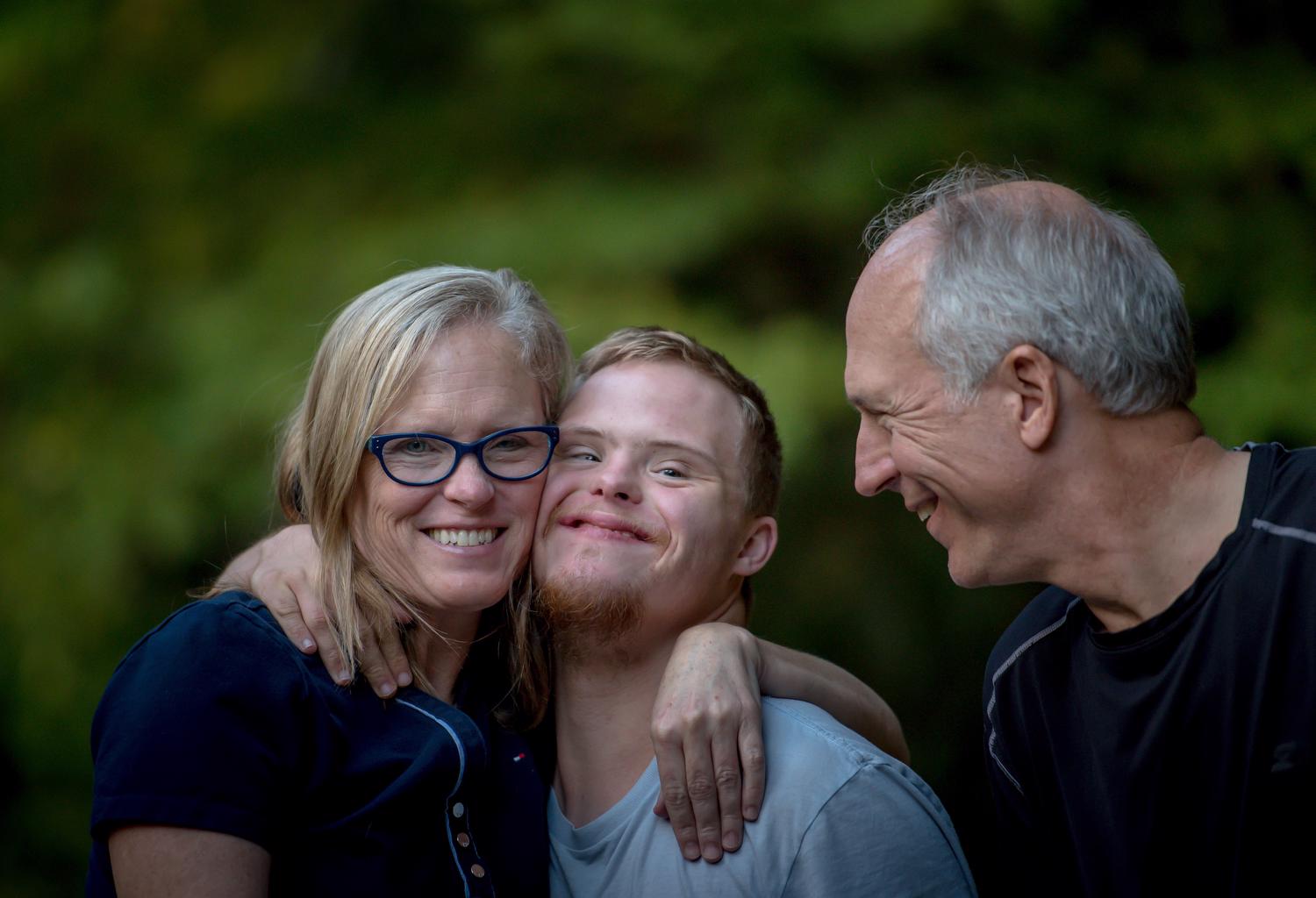 A family pose for a portrait. The mother has her arm around her son's shoulder and they are embracing cheek to cheek. The teenage son has down syndrome, and the father looks at the mother and son with a smile. 
