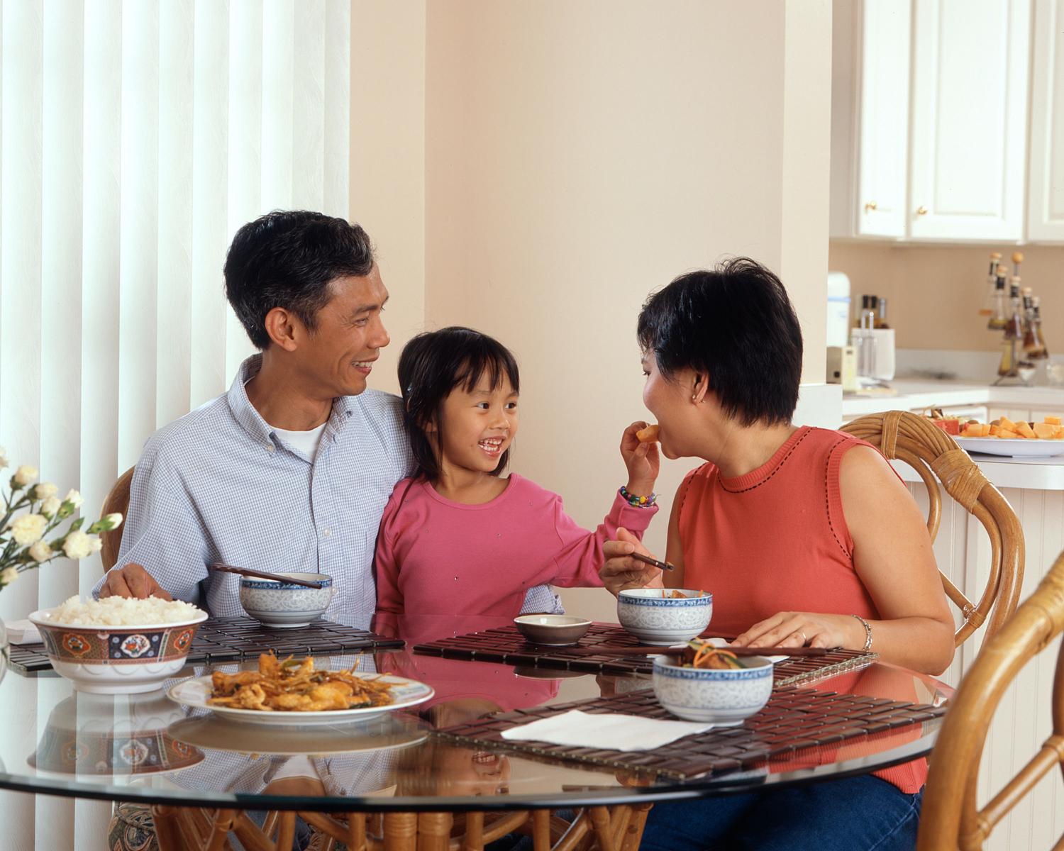 A young girl playfully feeds her mother a piece of food while sitting at the dinner table.