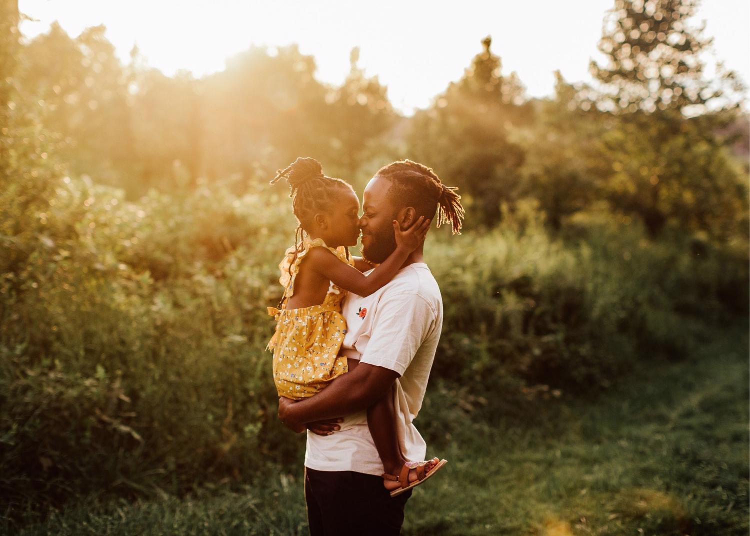 Man holding young girl; he is smiling while she rubs her nose against his outdoors with the sun behind