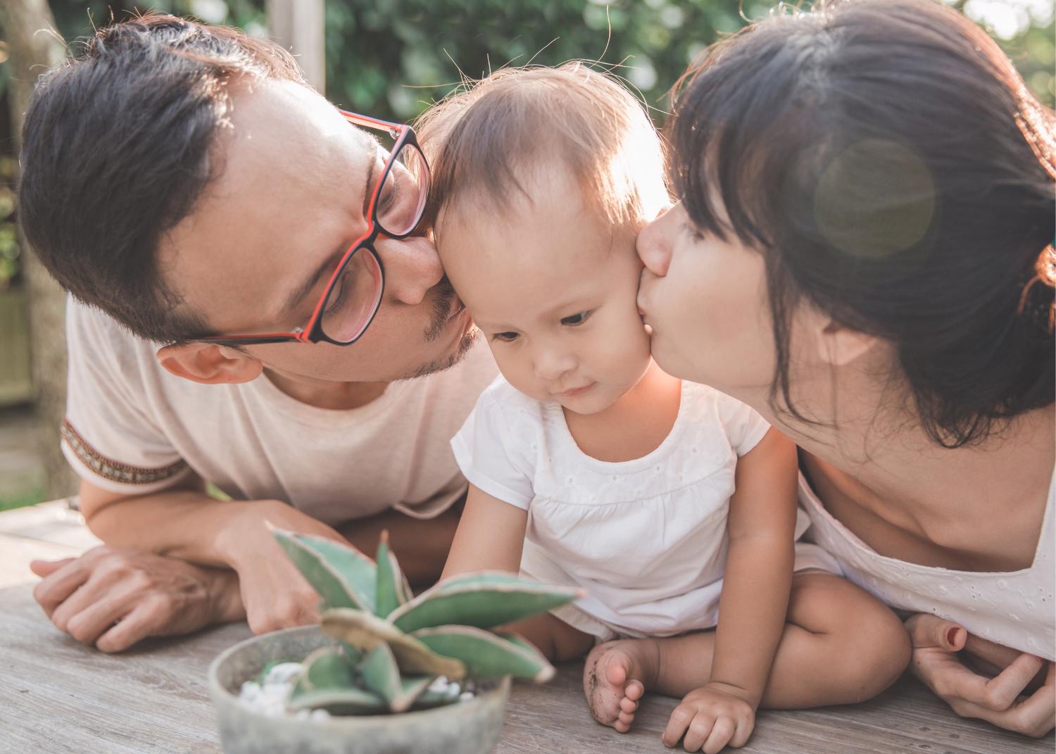 Small child looking at a plant, between a man wearing glasses and woman who are both leaned over kissing the child's cheeks
