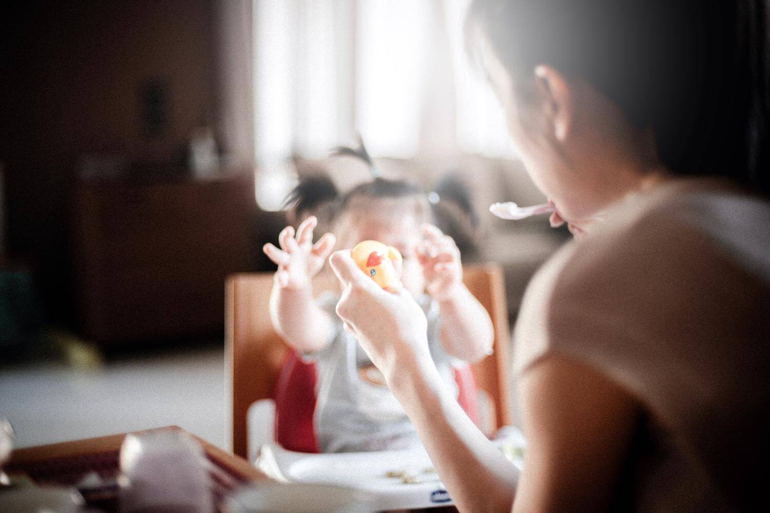 A toddler reaches for a toy as her female caregiver is holding a small spoon with food. 