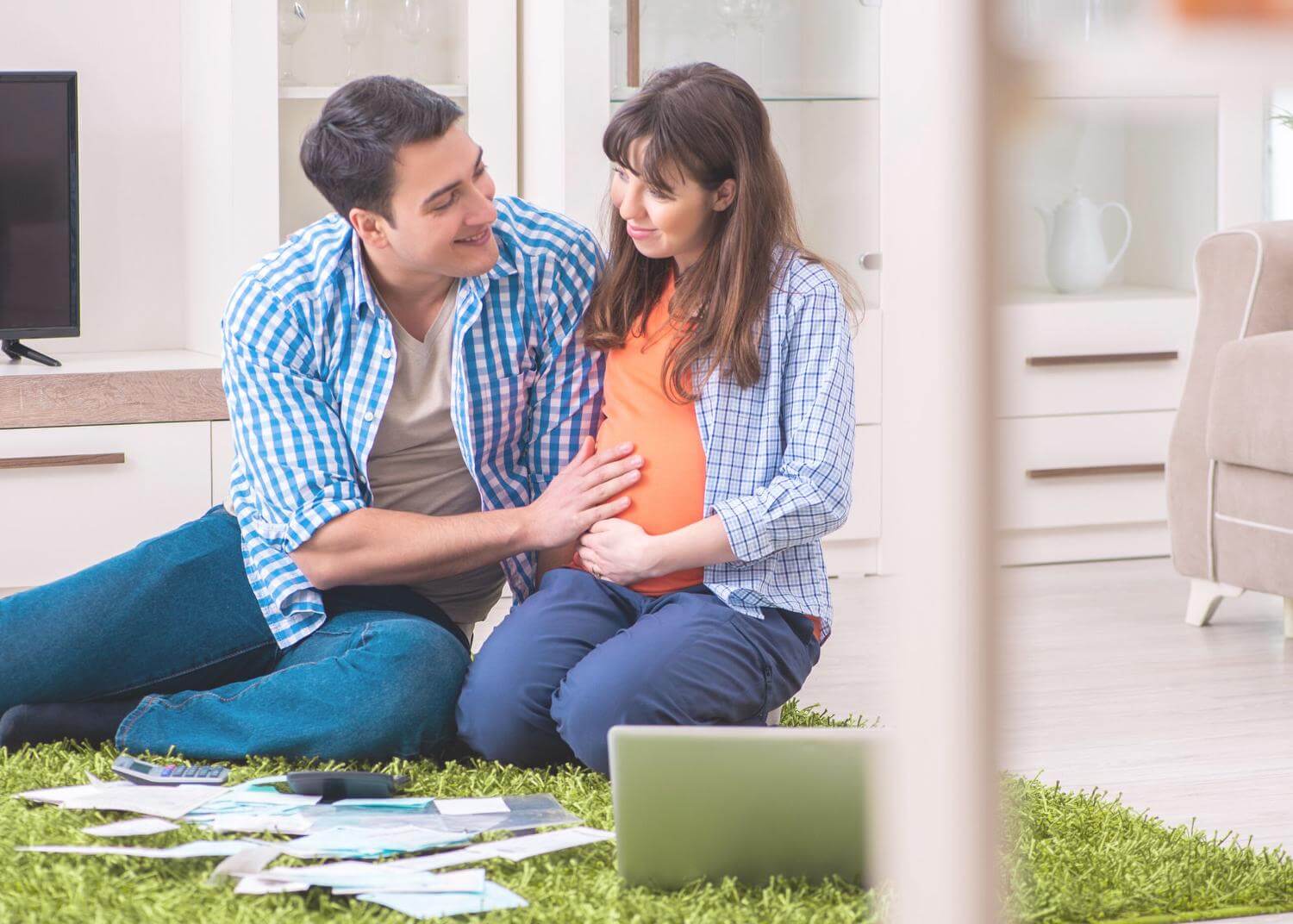 Man and woman sitting with bills and computer, smiling; woman is pregnant and man has his hand on her stomach