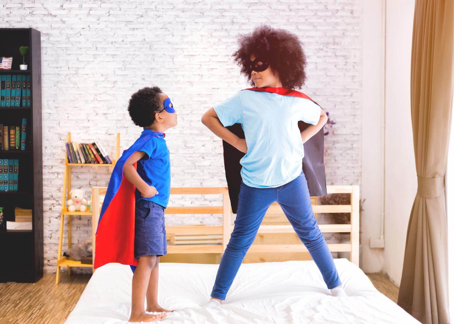 Two children standing on a bed wearing masks and capes, hands on hips