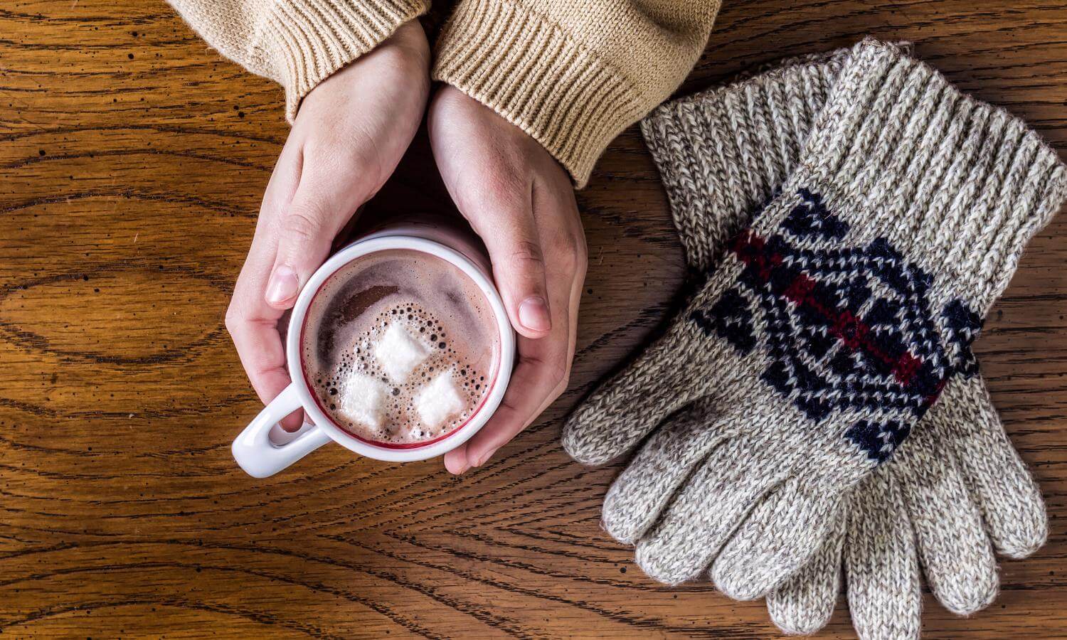 Hands holding mug of hot cocoa with marshmallows and gloves beside