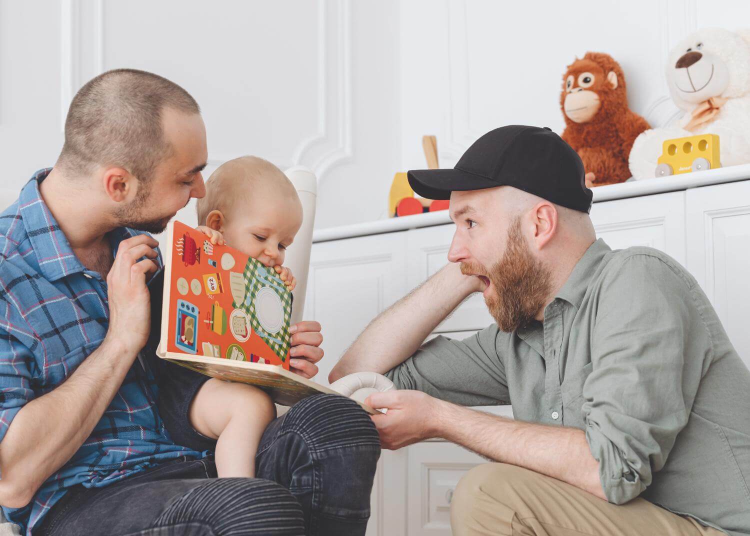 Two men smile and look on as a baby opens and peers into a book