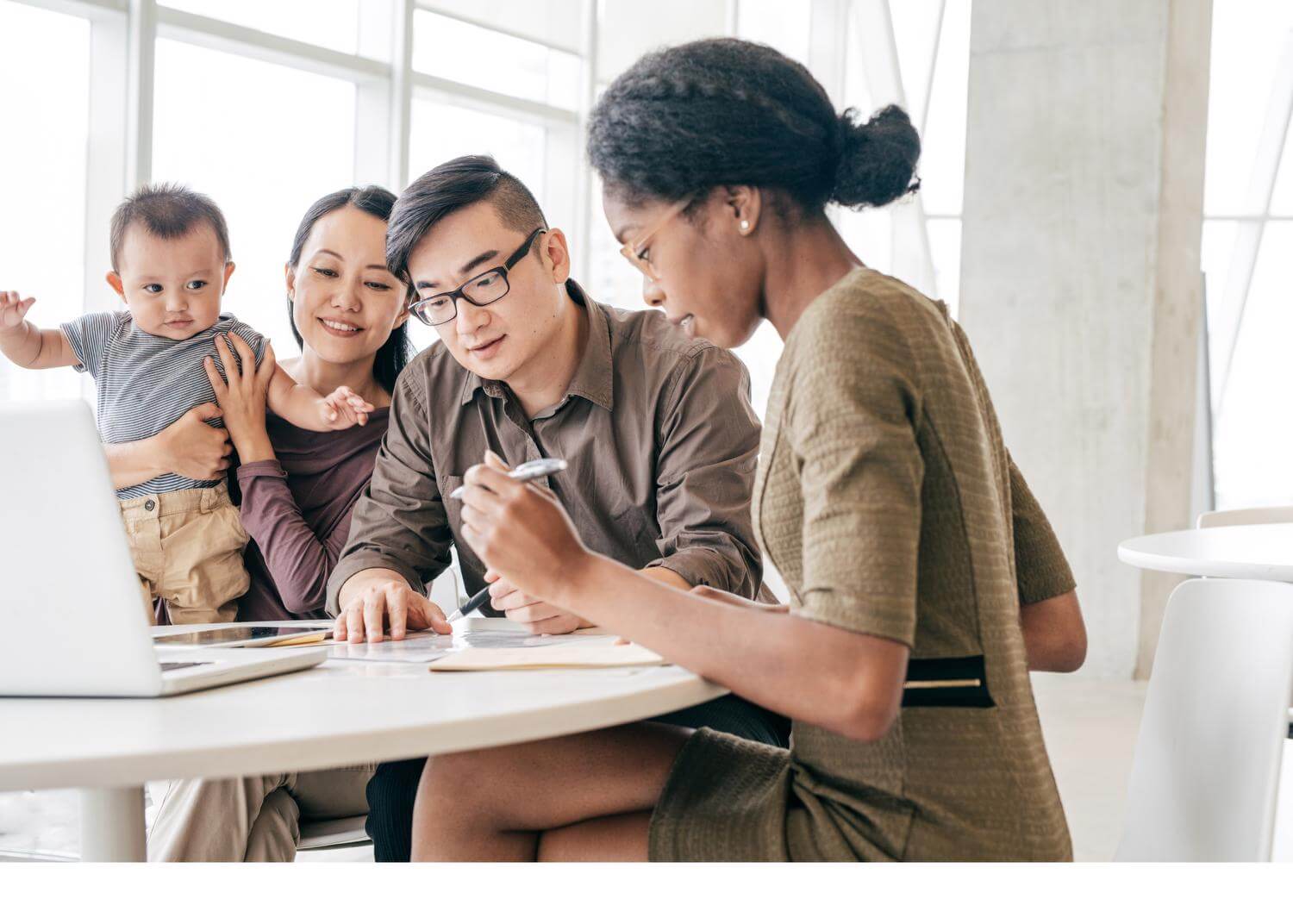 Woman holding toddler, man beside with additional woman holding pen/paper in front of computer