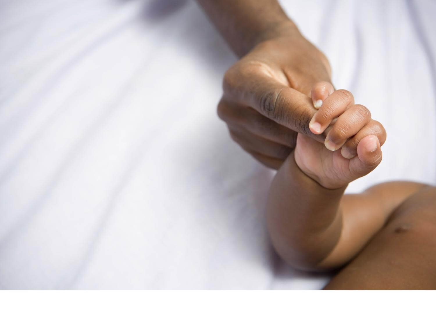 Infant hand holding an adult finger, both with brown skin