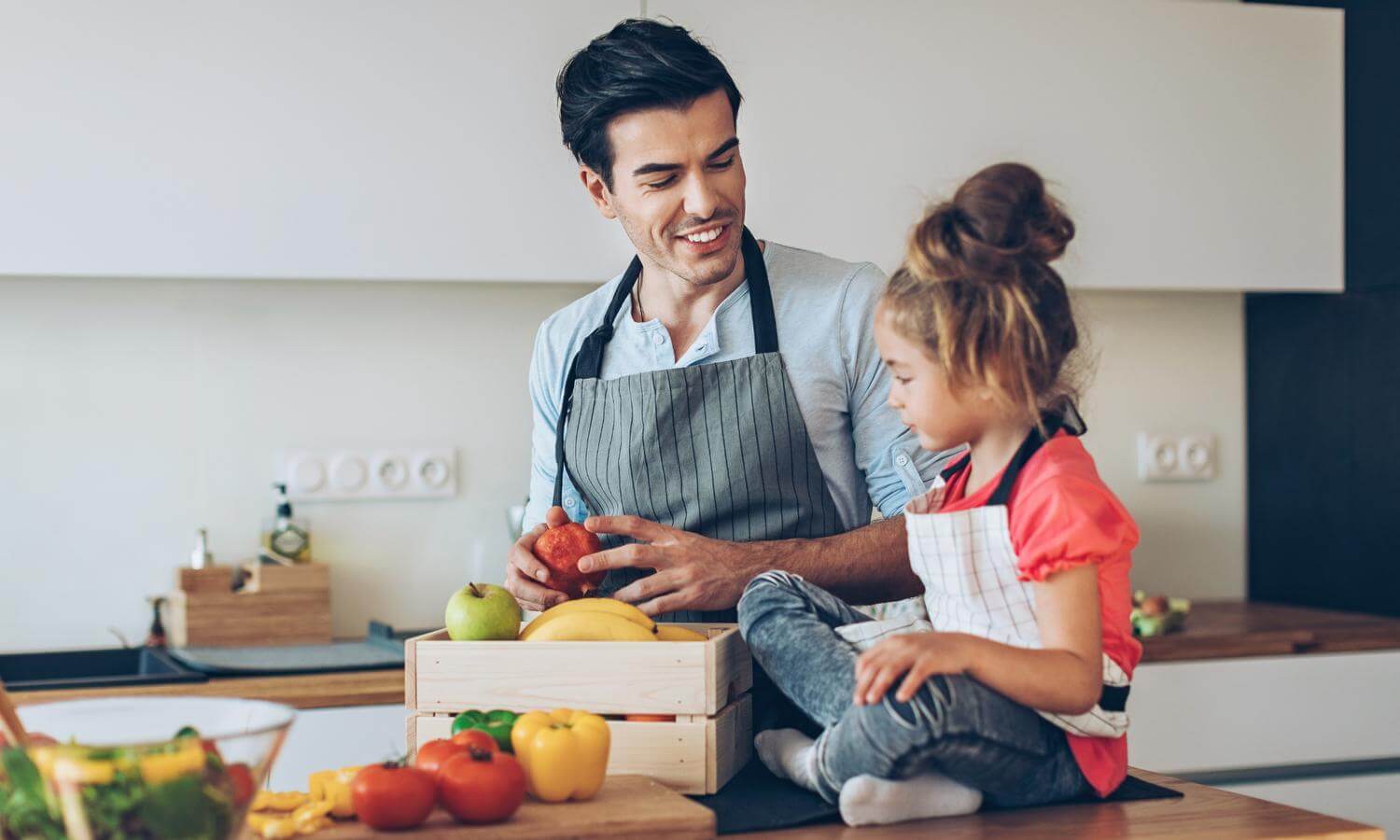 Adult father figure smiling at young girl sitting cross legged on counter with assortment of fruits and vegetables