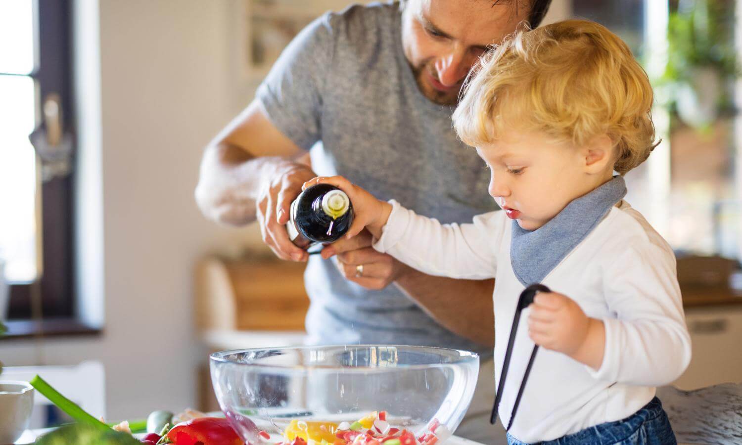 Father figure with young blonde child pour olive oil into bowl of vegetables