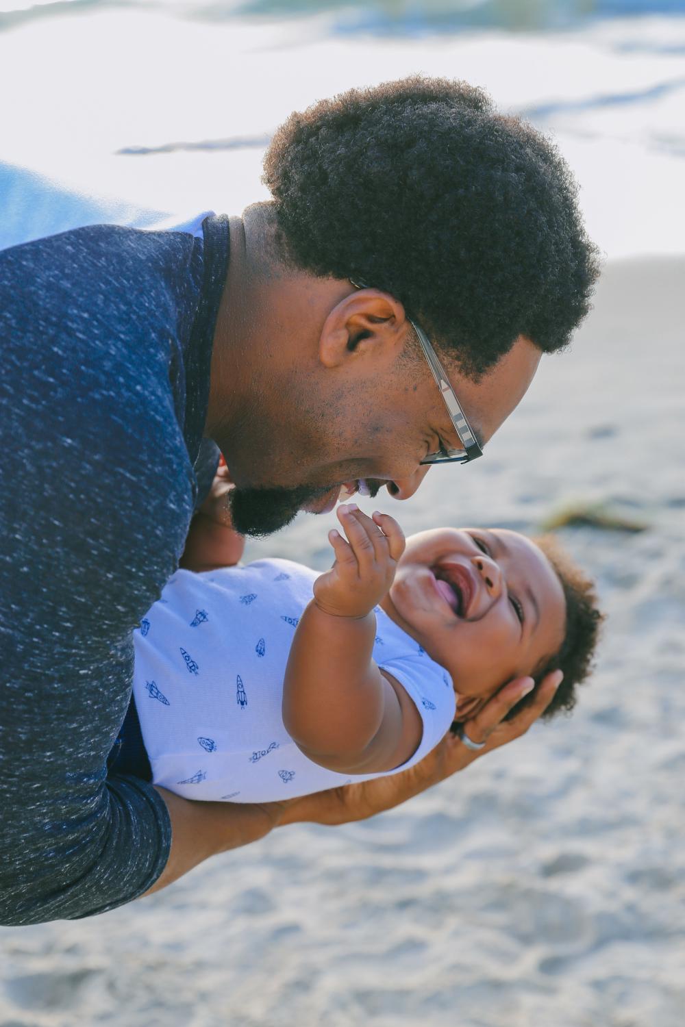 A Black father with glasses and a blue shirt playfully cradles his infant child while the infant laughs. They are on a beach.