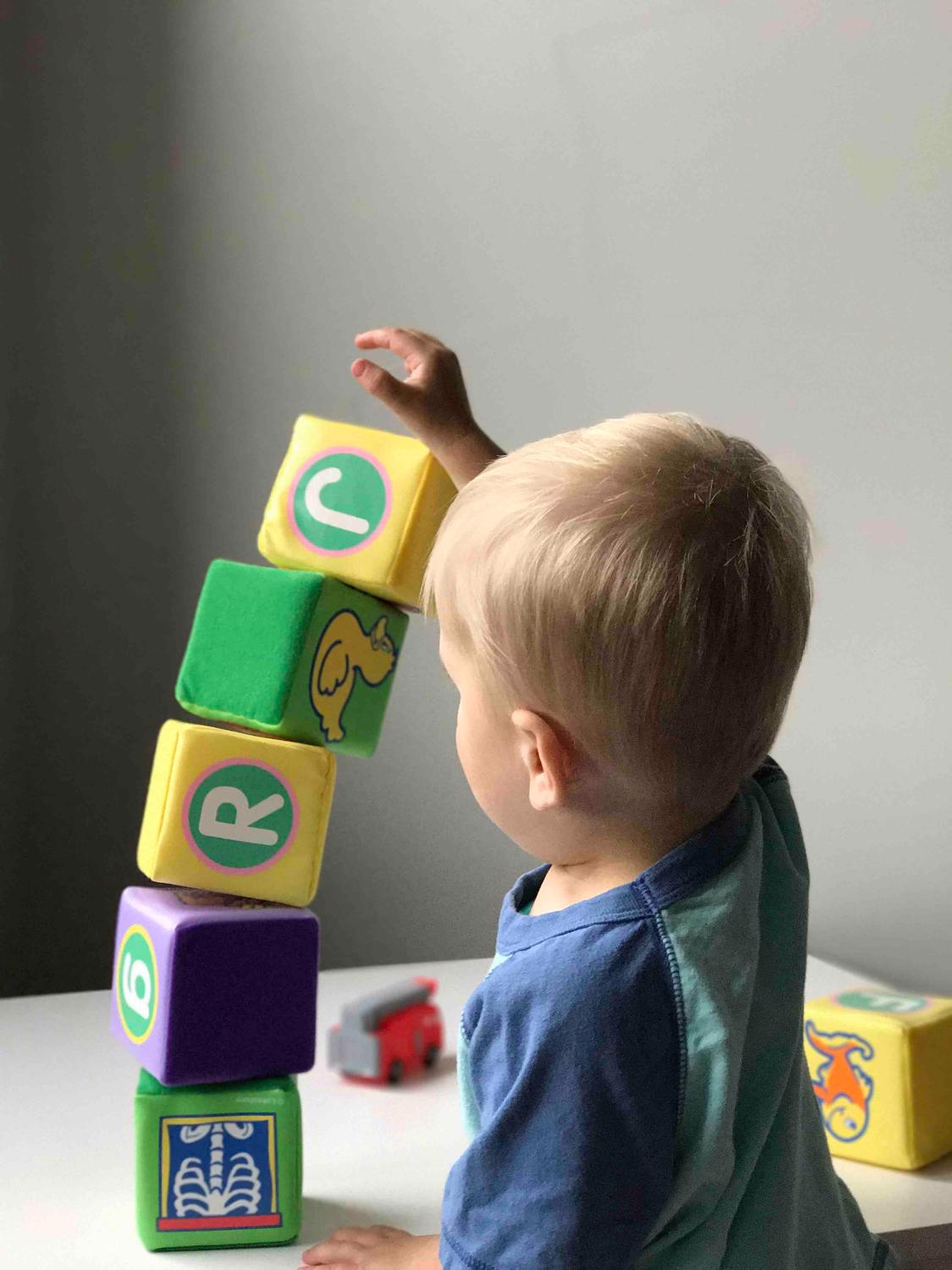 A toddler aged child with blonde hair stacks soft blocks on a table.