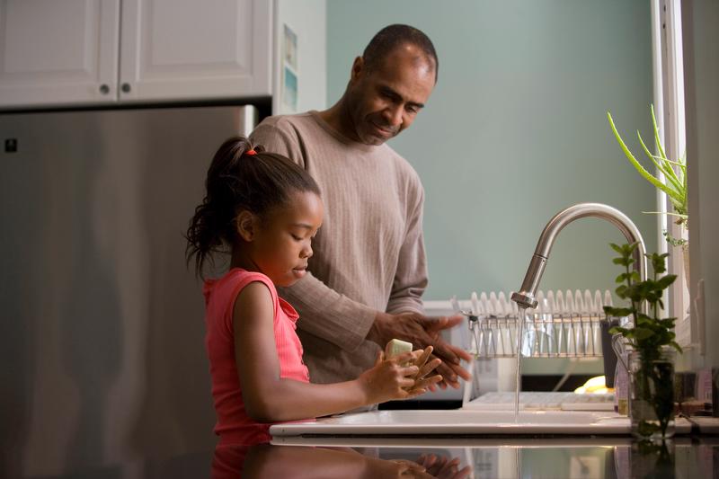 An adult, Black, male caregiver and a young Black girl are standing together at the kitchen sink. 