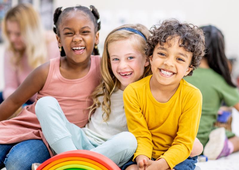 Three children sitting on the floor with arms around each other smiling and laughing.