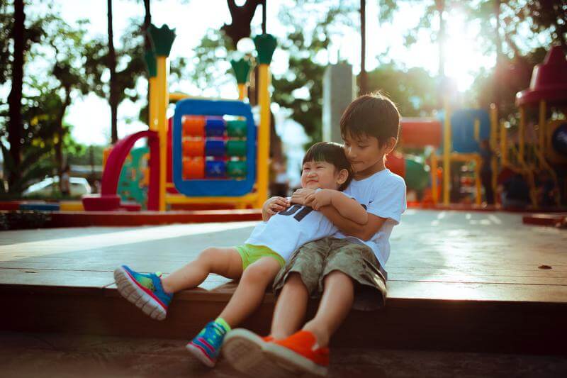 Two boys are playing on a playground. A boy around the age of seven with dark brown hair has his arms wrapped around a younger boys with short black hair who is leaning back against the chest of the older boy. They are both sitting and wearing shorts, t-shirts, and sneakers.