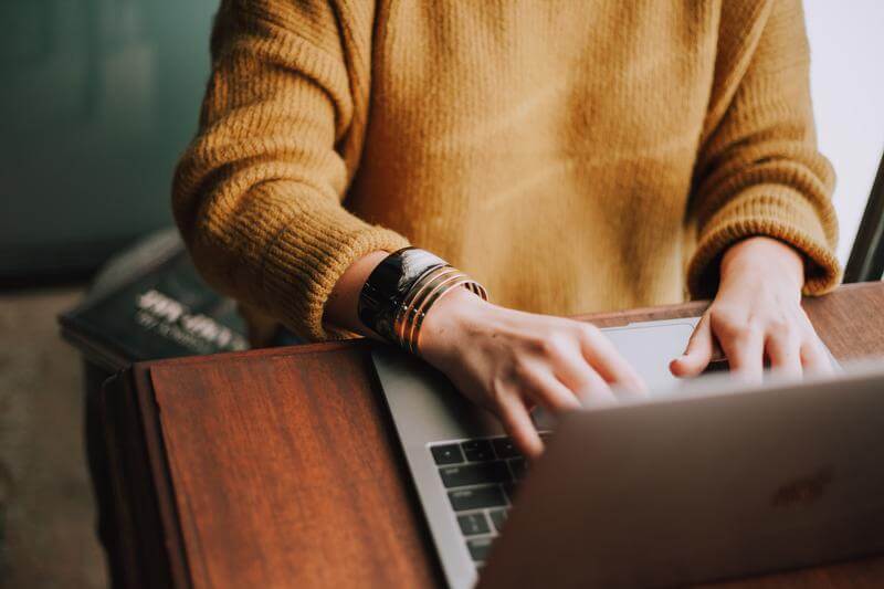 A close up of a woman in a mustard yellow sweater typing on a laptop. The image is cropped to only show the woman's arms, hands, and torso.